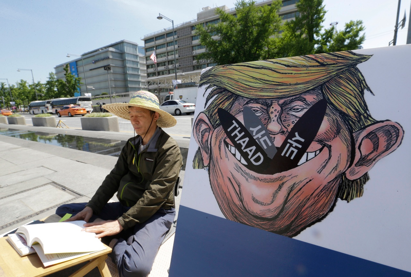 A South Korean protester sits next to a poster with an illustration of U.S. President Donald Trump to oppose a plan to deploy an advanced U.S. missile defense system called Terminal High-Altitude Area Defense, or THAAD, near the U.S. Embassy in Seoul on Monday, June 5, 2017 (AP Photo/Ahn Young-joon)